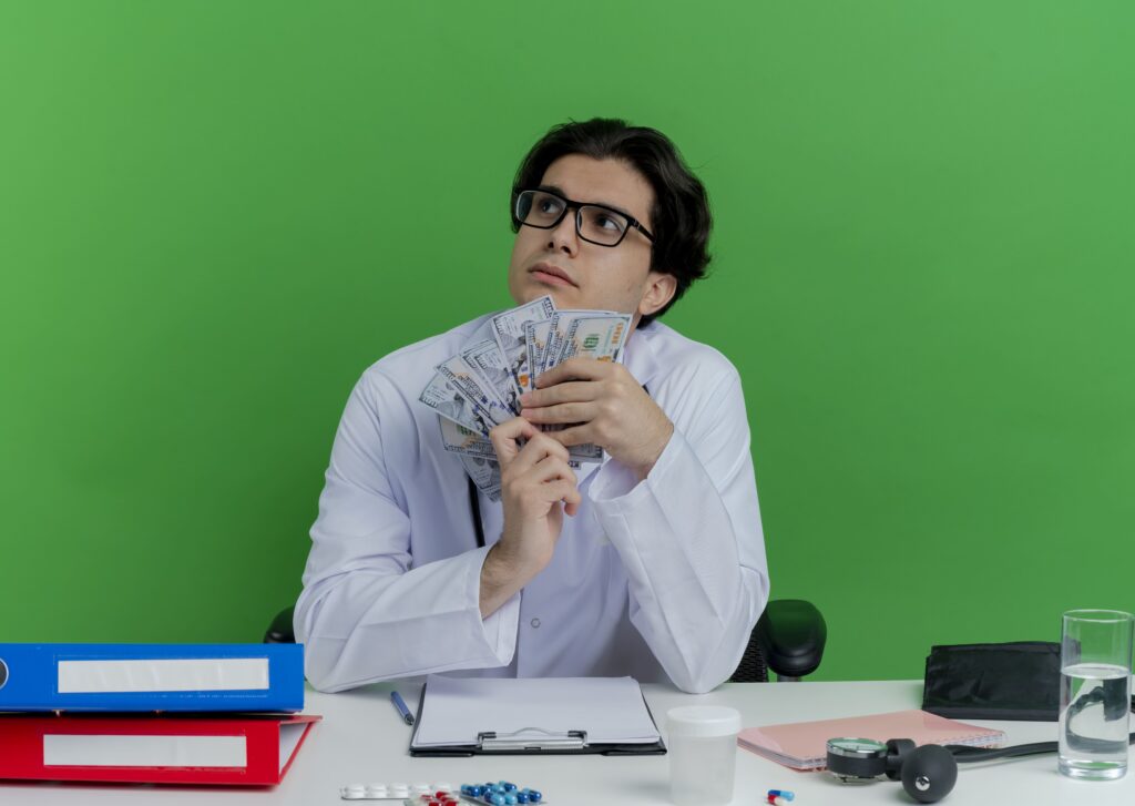 thoughtful-young-male-doctor-wearing-medical-robe-stethoscope-with-glasses-sitting-desk-with-medical-tools-holding-money-looking-side-isolated-green-wall