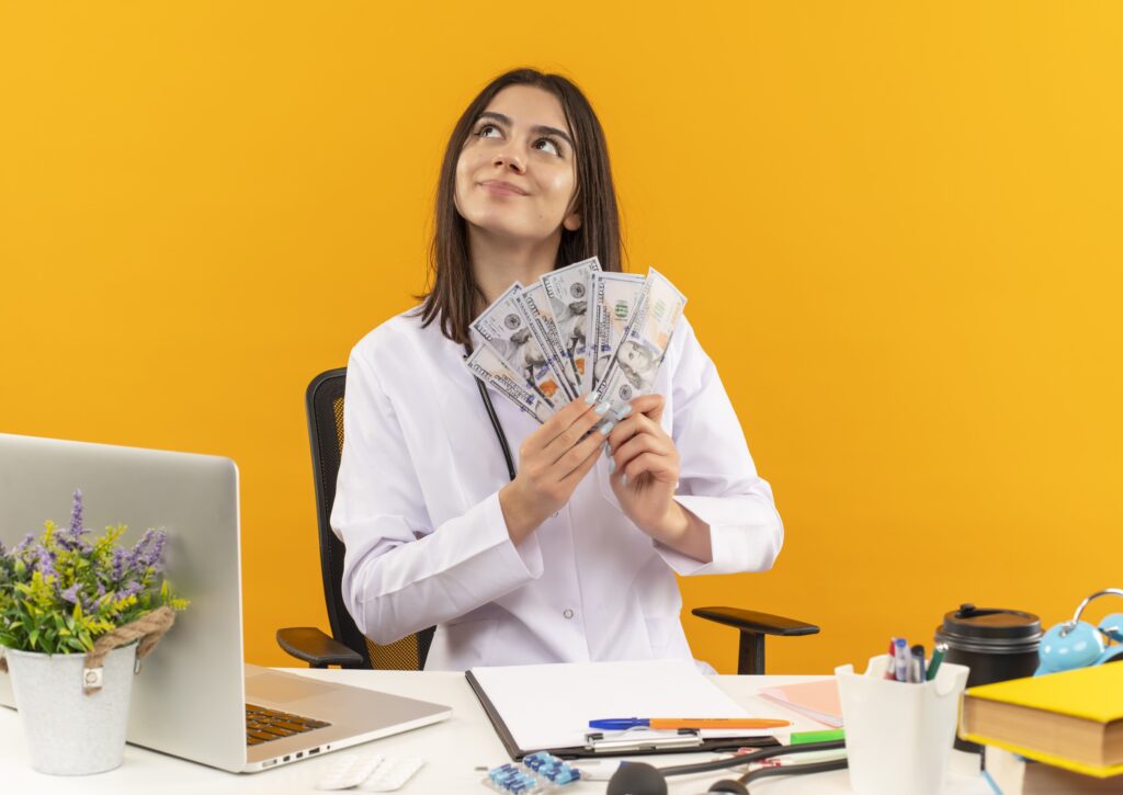 young-female-doctor-white-coat-with-stethoscope-holding-cash-with-dreamy-look-sitting-table-with-laptop-documents-orange-wall