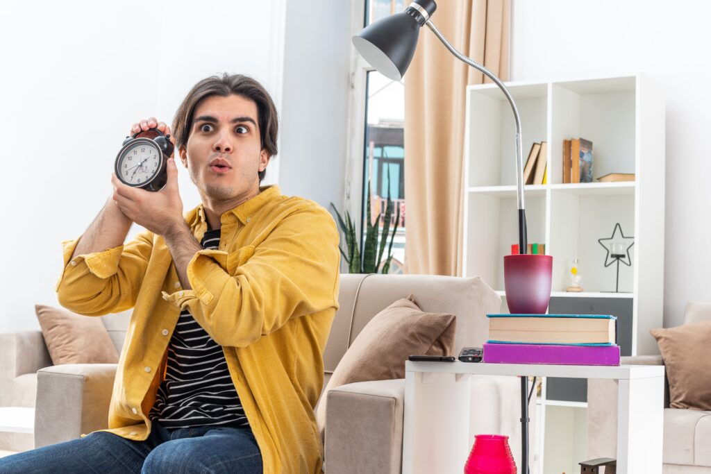 young-man-casual-clothes-holding-alarm-clock-looking-amazed-surprised-sitting-chair-light-living-room
